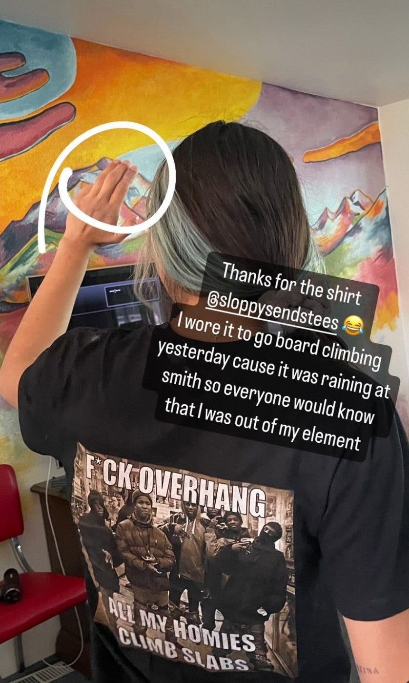 Rear view of a person wearing the black F*ck Overhang baby tee at a climbing gym, showing the All My Homies Climb Slabs print.